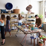 A bustling boutique shop with people browsing various items, including clothing and home decor, with a man organizing merchandise on a central table