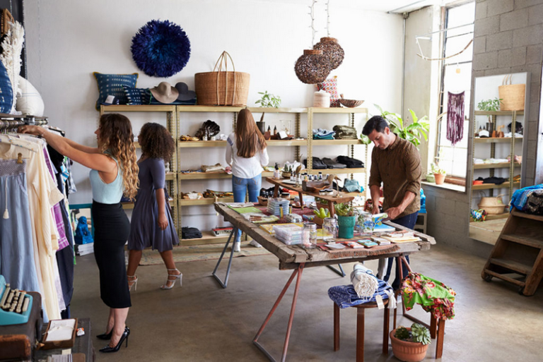 A bustling boutique shop with people browsing various items, including clothing and home decor, with a man organizing merchandise on a central table