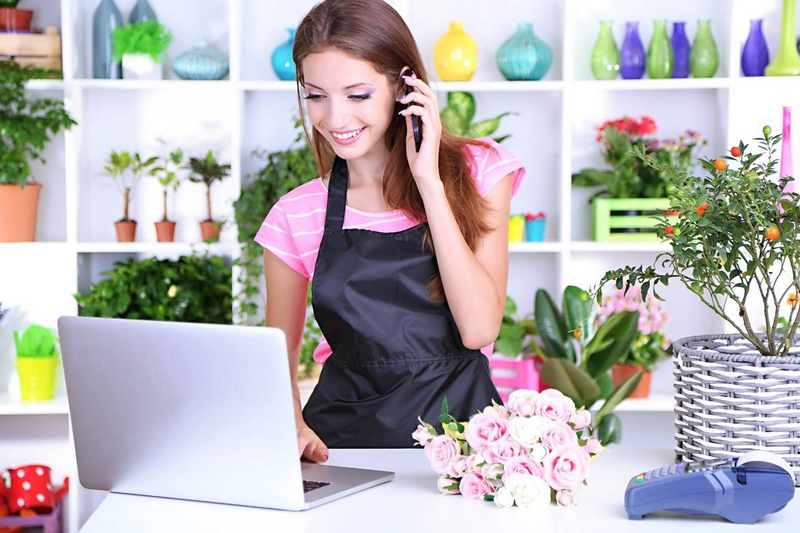 A woman in a black apron smiles while talking on the phone and using a laptop in a colorful room with shelves of decor